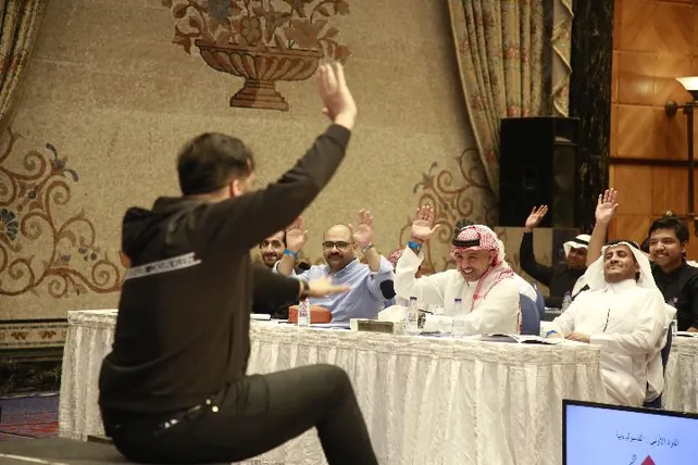 المدرّب جالسًا والحضور يرفعون أيديهم